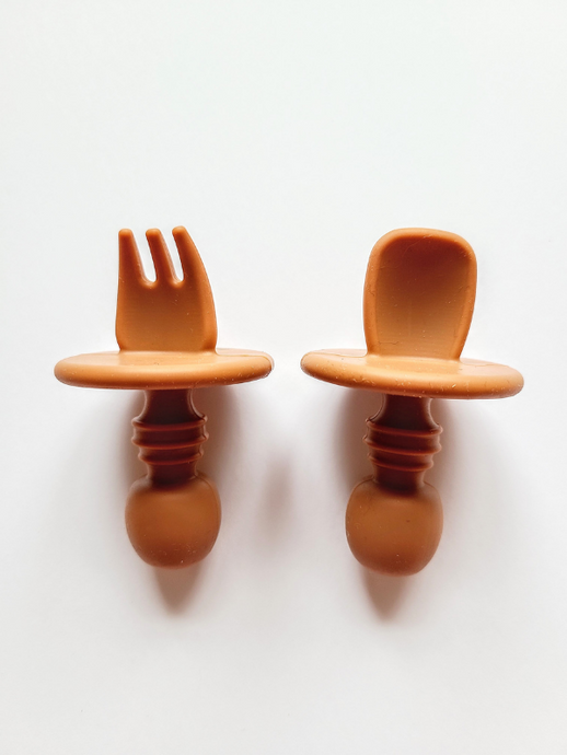 A burnt orange silicone spoon and fork utensil set meant for toddlers. The utensils are short and stubby, with a shield to prevent toddlers from inserting the utensil too far into their mouth and gagging on it.