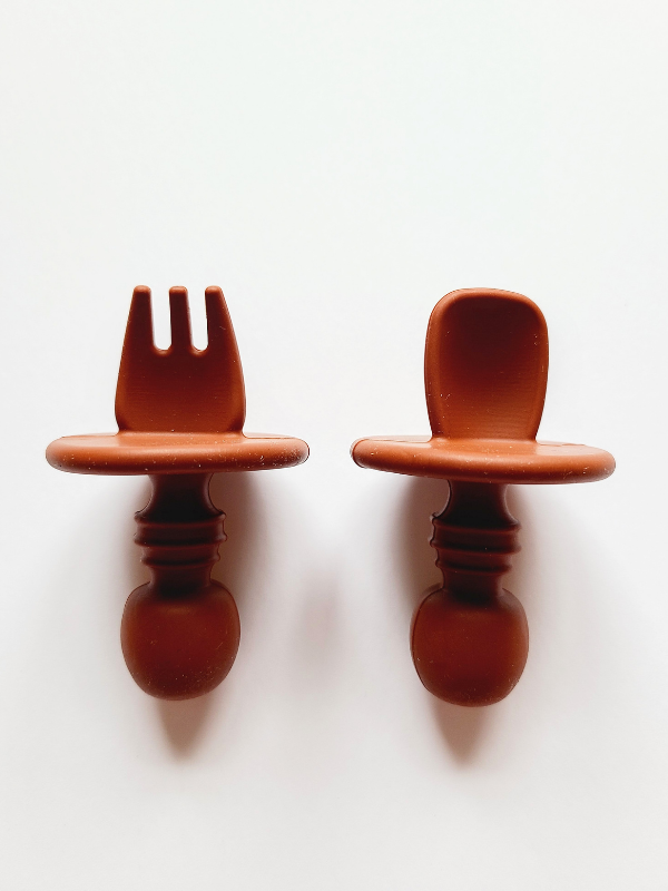 A clay brown silicone spoon and fork utensil set meant for toddlers. The utensils are short and stubby, with a shield to prevent toddlers from inserting the utensil too far into their mouth and gagging on it.