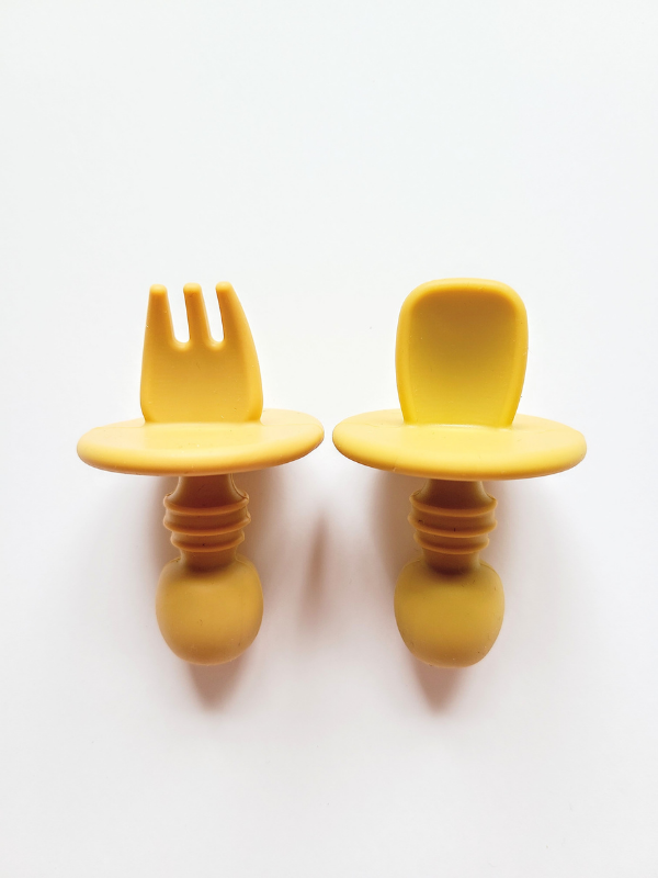A mustard yellow silicone spoon and fork utensil set meant for toddlers. The utensils are short and stubby, with a shield to prevent toddlers from inserting the utensil too far into their mouth and gagging on it.