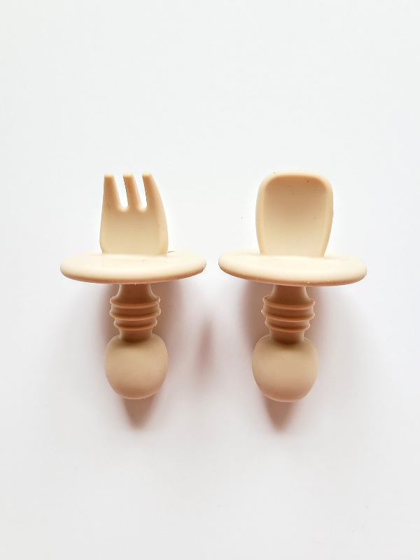A sand coloured silicone spoon and fork utensil set meant for toddlers. The utensils are short and stubby, with a shield to prevent toddlers from inserting the utensil too far into their mouth and gagging on it.  Edit alt text
