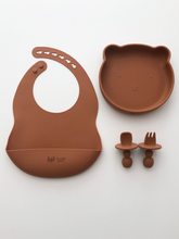 Load image into Gallery viewer, A silicone bib, bear plate, and baby utensil set (fork and spoon)  in a burnt orange colour. All the items are meant for use by babies or toddlers.
