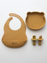 Load image into Gallery viewer, A silicone bib, bear plate, and baby utensil set (fork and spoon)  in a mustard yellow colour. All the items are meant for use by babies or toddlers.

