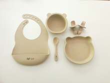 Load image into Gallery viewer, A silicone bib, spoon, bear plate, bear bowl, and baby utensils (fork and spoon) in a sand beige colour. All the items are meant for use by babies or toddlers.
