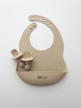 Load image into Gallery viewer, A silicone bib, and baby utensils (fork and spoon) in a beige sand colour. All the items are meant for use by babies or toddlers.
