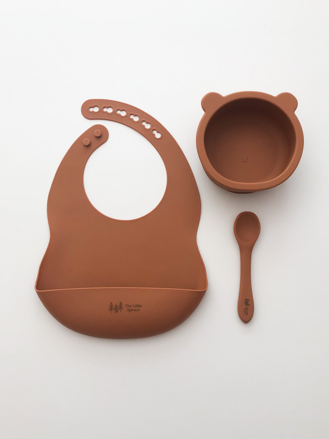 A silicone bib, bear bowl, and spoon in a burnt orange colour. All the items are meant for use by babies or toddlers.