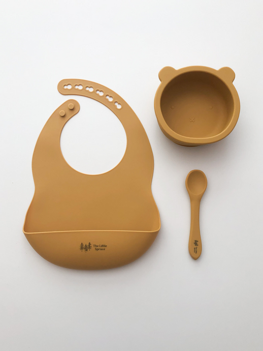 A silicone bib, bear bowl, and spoon in a mustard yellow colour. All the items are meant for use by babies or toddlers.